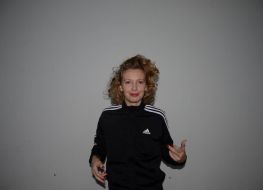 Na zdjęciu autorka warsztatów. Zdjęcie wykonane na szarym tle. Kobieta ubrana w czarno białą bluzę. Włosy ciemny blond, kręcone, półdługie. Uśmiechnięta. Zdjęcie do połowy tułowia.