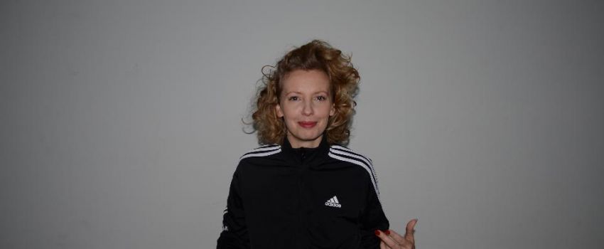 Na zdjęciu autorka warsztatów. Zdjęcie wykonane na szarym tle. Kobieta ubrana w czarno białą bluzę. Włosy ciemny blond, kręcone, półdługie. Uśmiechnięta. Zdjęcie do połowy tułowia.