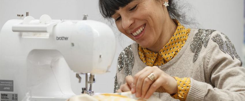 uśmiechnięta kobieta o ciemnych włosach, szyjąca na maszynie, na pierwszym planie wielobarwne tkaniny