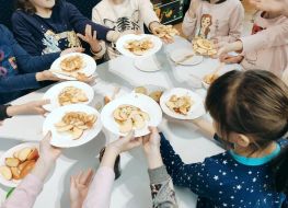 Wokół stołu grupa dzieci. W wyciagniętych dłoniach trzymaja talerze, na nich pokrojone w plasterki jabłka