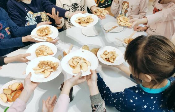 Wokół stołu grupa dzieci. W wyciagniętych dłoniach trzymaja talerze, na nich pokrojone w plasterki jabłka