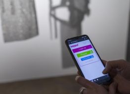 Na pierwszym planie dłonie trzymające smartfon. W tle srebrny obiekt