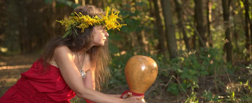 Kobieta w czerwonej sukience i wiankiem z żółtych kwiatów siedzi na leśnej drodze prawym bokiem do widza. W wyciągniętych dłoniach trzyma instrument muzyczny Ipu. W tle las.