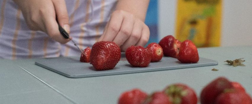 na zdjęciu lśniące świeże truskawki. Ktoś kroi je nożem na szarej desce