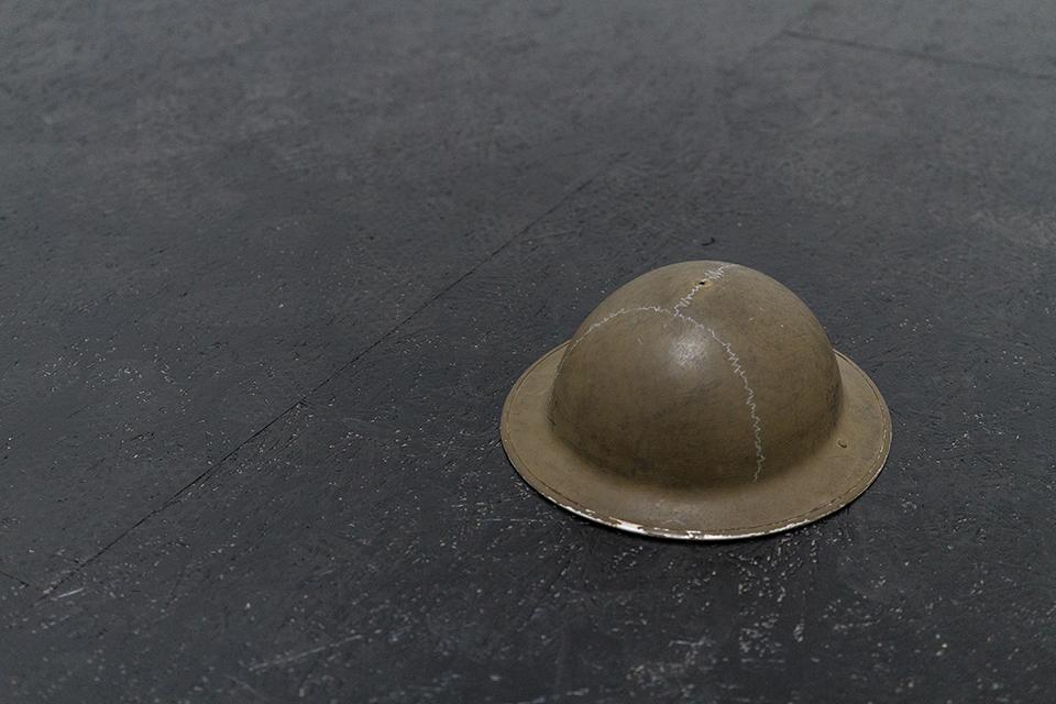 Hełm w odcieniu brudnej zieleni. Leży na ciemnej podłodze po prawej stronie zdjęcia.