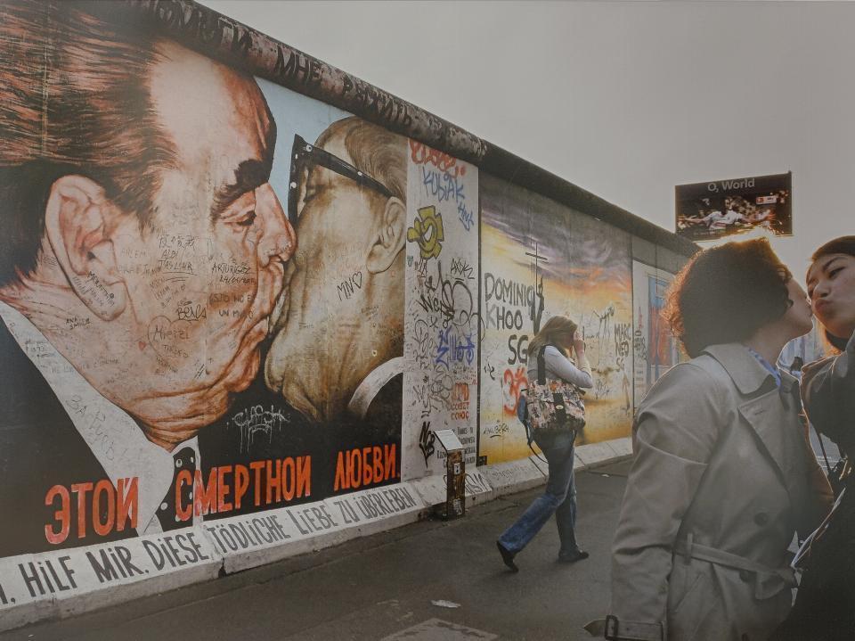 Zdjęcie przedstawia dwie młode kobiety na tle muru berlińskiego pokrytego graffiti.