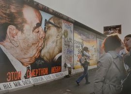 Zdjęcie przedstawia dwie młode kobiety na tle muru berlińskiego pokrytego graffiti.