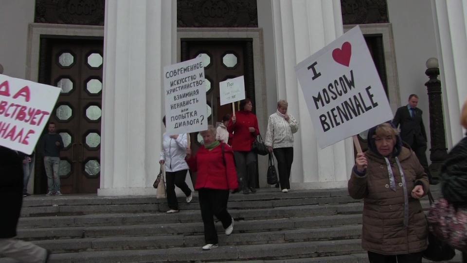 elderly women on the stairs in front of the building, holding banners in Russian and English in their hands