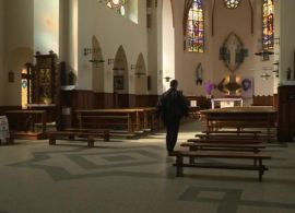 Widok wnętrza świątyni. Po obu stronach rzędy ławek. Pomiędzy nimi ubrany na czarno mężczyzna. Widać jego tył.