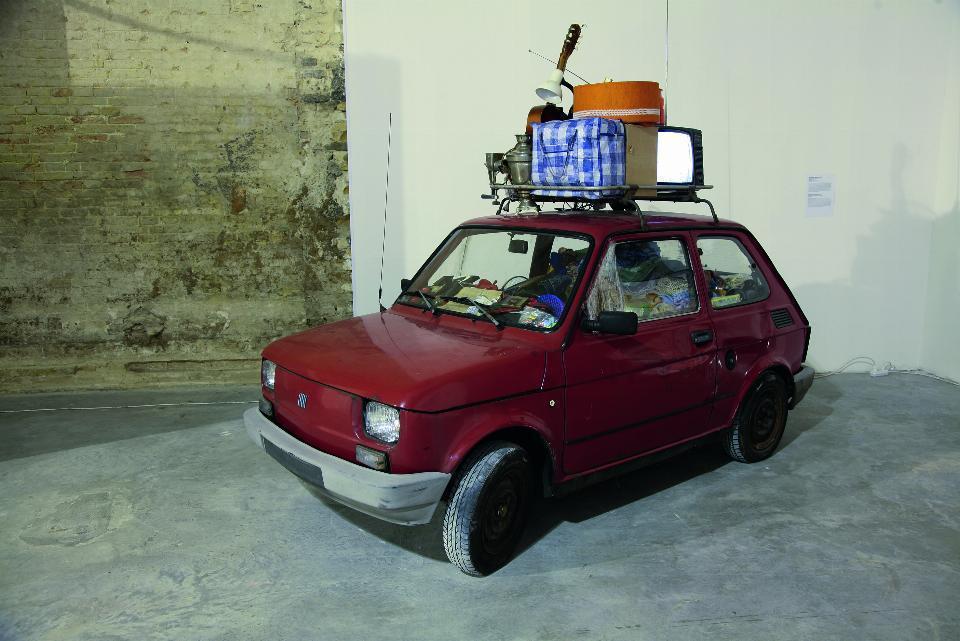 Ciemoczerwony mały fiat 126p. Zagracony w środku. Na dachu umieszczone są bagaże. Stoi bokiem w pomieszczenu z betonową podłogą i białymi ścianami. Fragment ściany po lewej stronie z widoczną cegłą.