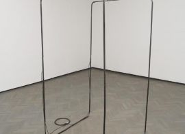 Metalowa konstrukcja zblizona do prostopadłościanu. Po lewej kabel zwinięty w pętle. Stoi na drewnianej podłodze na tle białej ściany.