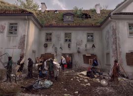 Zniszczony budynek z dachem porośniętym mchem i roślinami. Przed nim grupa ludzi w zniszczonych ubraniach. Wokół panuje bałagan.