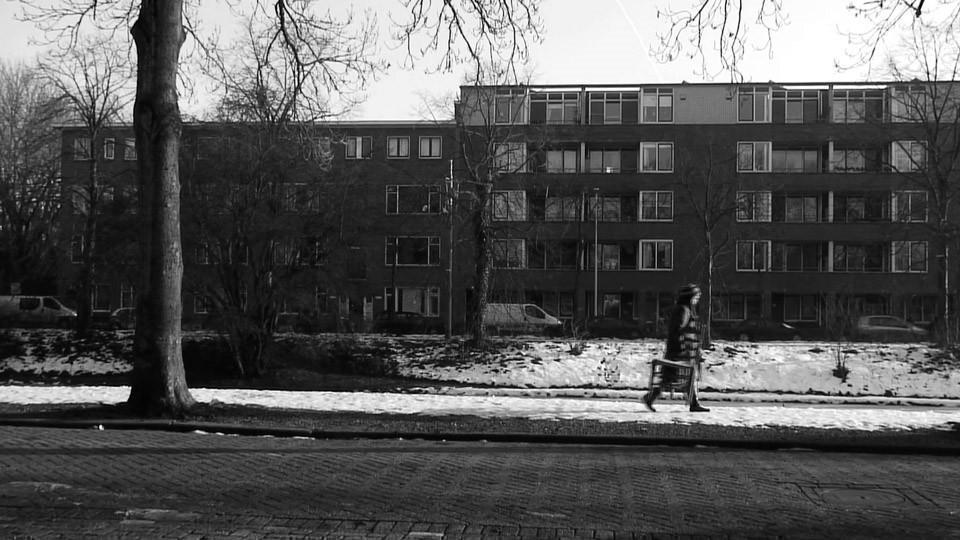 Czarnobiała fotografia przedstawiająca fragment osiedla w zimie. Po prawej stronie widać postać idącą po nieodśnieżonej ścieżce. Jest nieostra. W prawej dłoni niesie przedmiot. Po lewej stronie znajduje się fragment pnia drzewa. W tle niski blok.