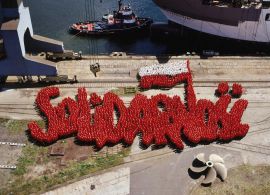 1 część dyptyku. Zdjęcie przedstawia kilka tysięcy żołnierzy, ubranych  w czerwone i białe bluzy, ustawionych w taki sposób, że tworzą znak Solidarności. Dominuje czerwień, biel pojawia się w polskiej fladze. Stoją na betonowym nabrzeżu.