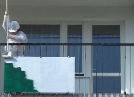 Widok na balkon. Na nim znajduje się osoba w białym ubraniu ochronnym. Maluje na biało płytę umieszczoną na zewnętrznej stronie balustrady, po lewej. Pod białą farbą widoczny zielony fragment.