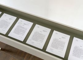 Leżące obok siebie białe kartki z wydrukowanym podaniem. Umieszczone są w białej gablocie za szkłem. W tle widoczny fragment drewnianej podłogi i białej ściany.