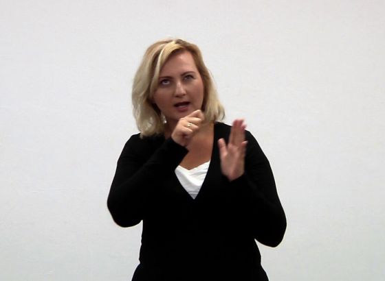 Młoda kobieta z jasnymi włosami ubrana w białą podkoszulkę i czarny sweter. Pracuje dłońmi. To tłumaczka języka migowego