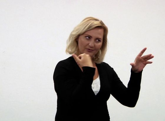 Młoda kobieta z jasnymi włosami ubrana w białą podkoszulkę i czarny sweter. Pracuje dłońmi. To tłumaczka języka migowego