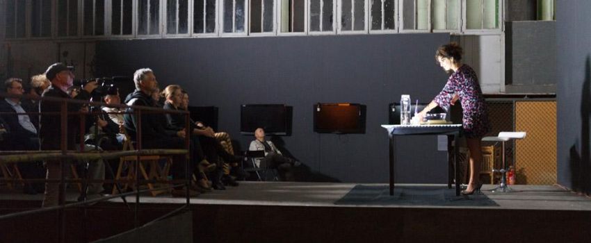 Zdjęcie wykonane w przestrzeni Galerii Arsenał elektrownia. W pomieszczeniu znajduje się grupa osób siedzacych na krzesełkach. Naprzeciwko ich stoi kobieta pochylająca się nad stolikiem. Tło stanowi szara ściana z dużą ilością okiennic.