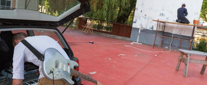 Zdjęcie przedstawia plac przed budynkiem Galerii Arsenał elektrownia. Mężczyzna ubrany w białą bluzkę i brązowe spodnie pochyla się nad bagażnikiem samochodu. Na plecach zawieszoną ma gitarę elektryczną. Druga osoba znajduje się w oddali. Siedzi ona na wy