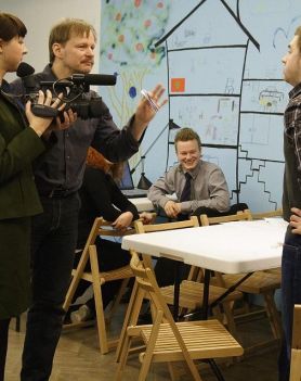 Na zdjęciu widoczna grupa osób - dwie kobiety i trzech mężczyzn. Jedna z kobiet trzyma w dłoniach kamerę. Obok nich znajdują się stoliki, krzesła. W tle widoczny telewizor oraz błękitno-białe ściany pomieszczenia.