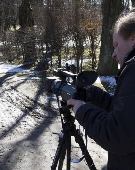 Na zdjęciu widoczny jest ubrany w ciemną kurtkę mężczyzna znajdujący się na leśnej drodze. Dotyka on kamery ustawionej na statywie. W tle widoczne drzewa.