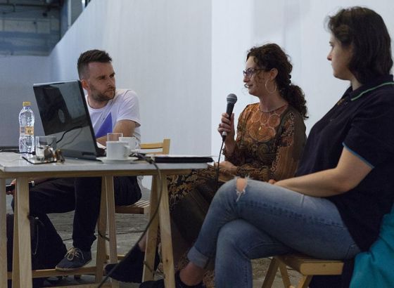 Dwie kobiety i mężczyzna siedzą przystoliku. Jedna z kobiet trzyma w dłoni mikrofon. Na stole znajduje się laptop, stoją filiżanki oraz butelka z wodą