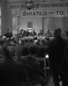 Stara fotografia. Na krzesłach siedzą ludzie. W niewielkiej odległości, przed nimi stoi długi, prostokątny stół przy którym ównież siedzą ludzie. Za ich plecami wisi tranparent z napisem ŚWIAŁO TO SIŁA".  Zdjęcie czarno-białe.