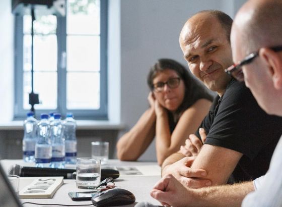 Dwóch mężczyzn i kobieta siedzą przy stole. Postać kobiety mniej wyraźna. Na stoliku stoją butelki z wodą, szkalnki, leży mikrofon. Zdjęcie zrobione z bocznej perspektywy.