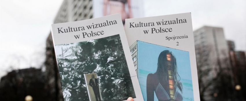 Na pierwszym planie dłoń trzymająca dwie książki. W tle blokowisko.