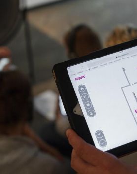 Dłoń trzymająca tablet. W tle ludzie siedzący na podłodze. Tło rozmyte.