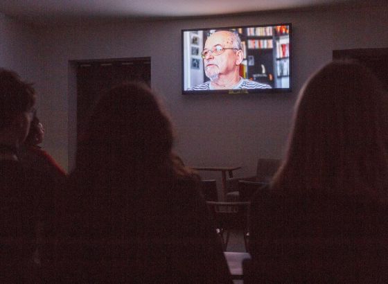 Ma pierwszym planie widać plecy siedzących dwóch kobiet. W dalszym zaś widoczny jest wiszący na białej ścianie telewizor, na którym wyświetlany jest obraz. Poniżej, przy ścianie widać fragment stojącego stolika i fotela. W pomieszczeniu jest zgaszone świa