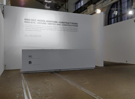 Wnętrze starej hali elektrowni Galerii Arsenał. Widok białej ściany na której przyklejone są białe napisy. Przed ścianą stoi  biurko z ladą.