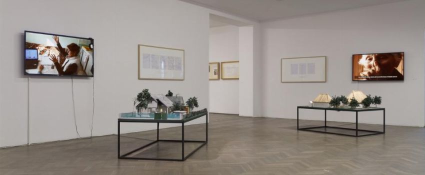 Zdjęcie wnętrza pomieszczenia. Na białych ścianach wiszą obrazy i telewizory z wyświetlanym obrazem. Na środku pomieszczenia stoją dwa stoły na których znajdują się instalacje.