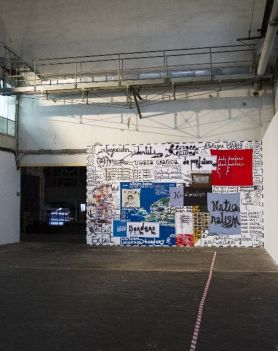 Widok ogólny wystawy w hali elektrowni Galerii Arsenał. Duże pomieszczenie, po obu stronach białe ścianki. Czołowa ściana wyklejona plakatami.