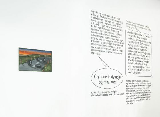 Fragment białej ściany. Po lewej stronie jest zawieszony obrazek, po prawej tekst opisujący wystawę.