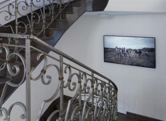 Projekcja wideo wyświetlana na telewizorze na białej ścianie. Po lewej stronie widoczny fragment schodów z balustradą.